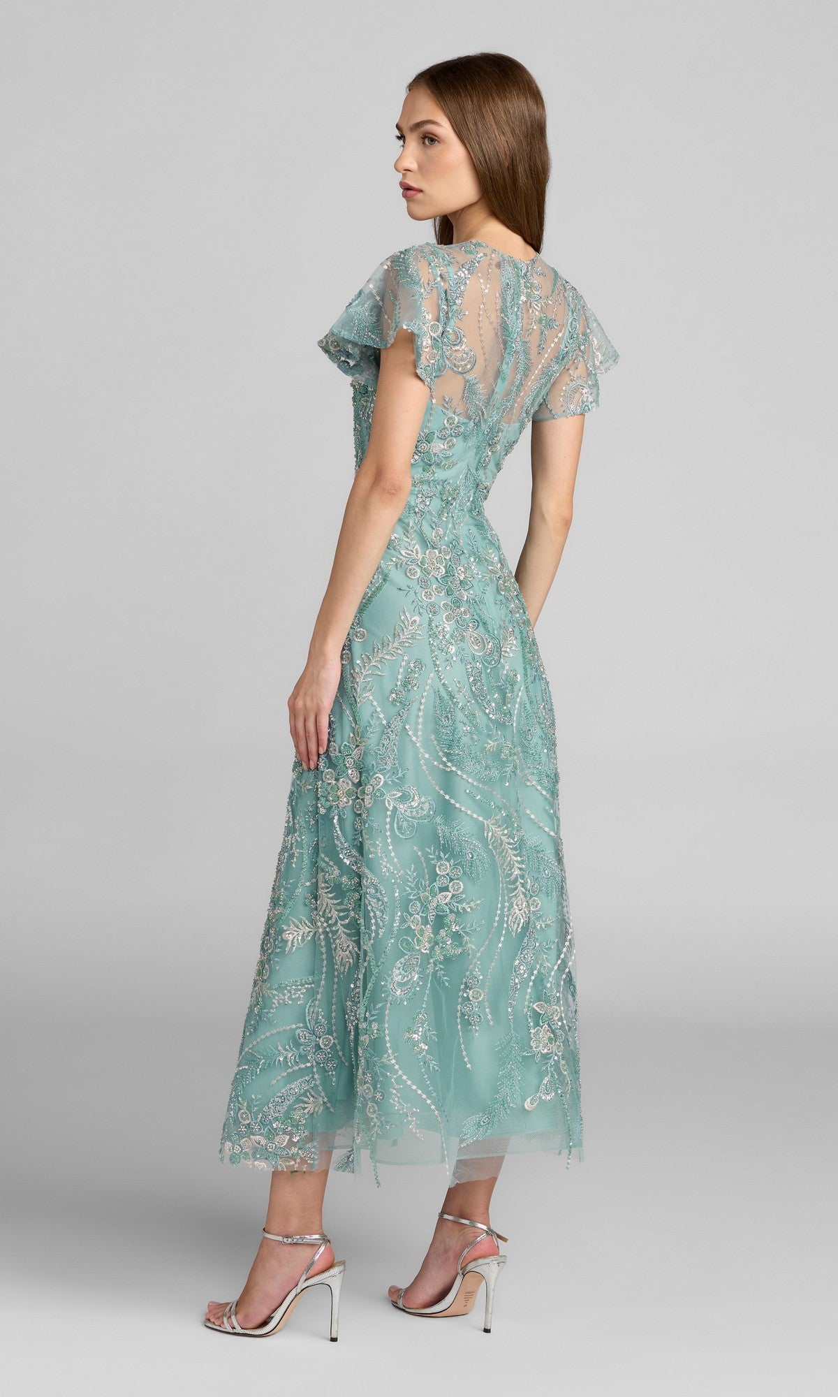 A woman stands sideways against a plain gray background wearing the Tea-Length Party Dress: Mac Duggal 68909—a light blue-green midi dress adorned with intricate beaded embroidery and sheer short sleeves—paired with silver high-heeled sandals.